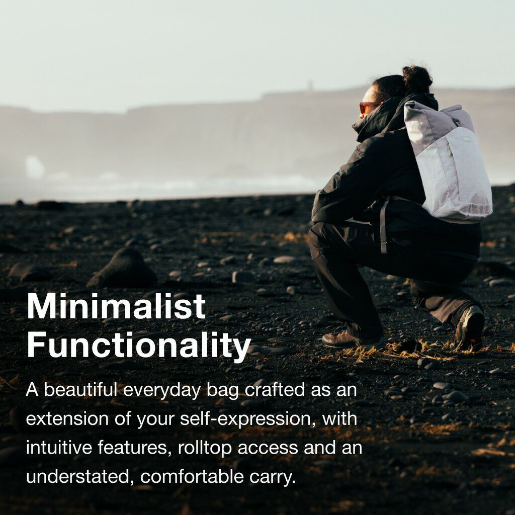 The image features a person crouched on a rocky black sand beach, wearing a dark jacket and pants. They have a white rolltop backpack on their back. The beach has scattered small rocks and the background shows a misty, distant landform under a pale sky. T