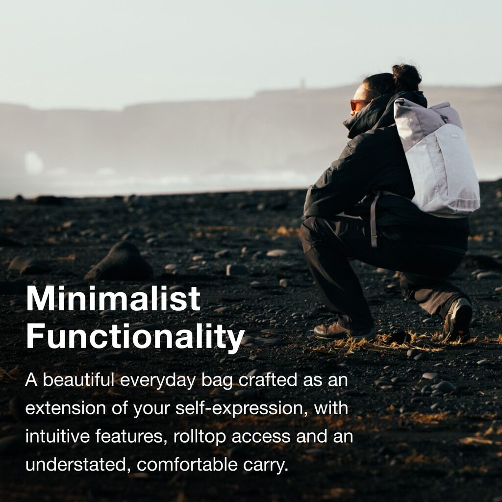 The image features a person crouched on a rocky black sand beach, wearing a dark jacket and pants. They have a white rolltop backpack on their back. The beach has scattered small rocks and the background shows a misty, distant landform under a pale sky. T