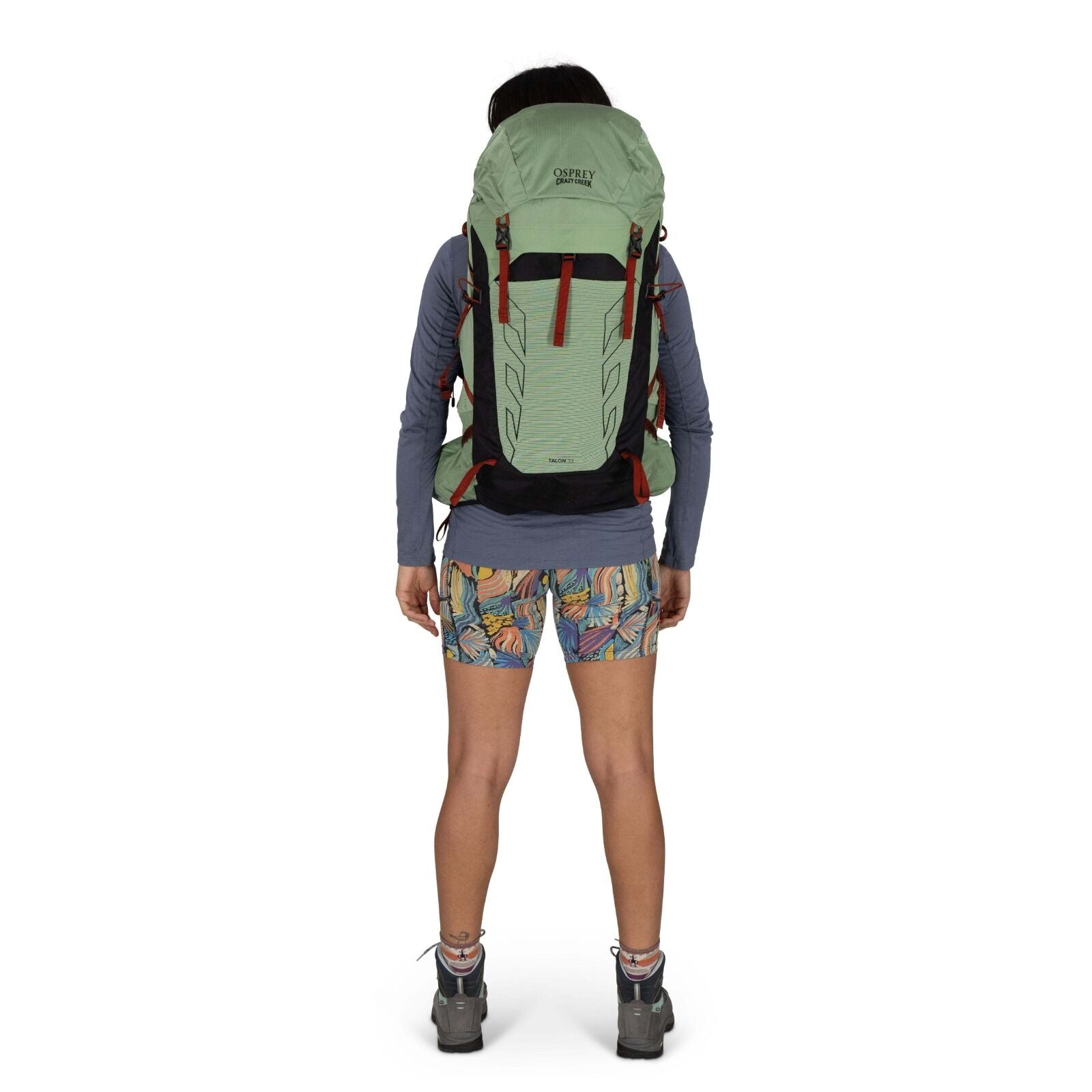 A woman stands with her back to the camera, wearing a green Osprey backpack and colorful patterned shorts, ready for adventure.