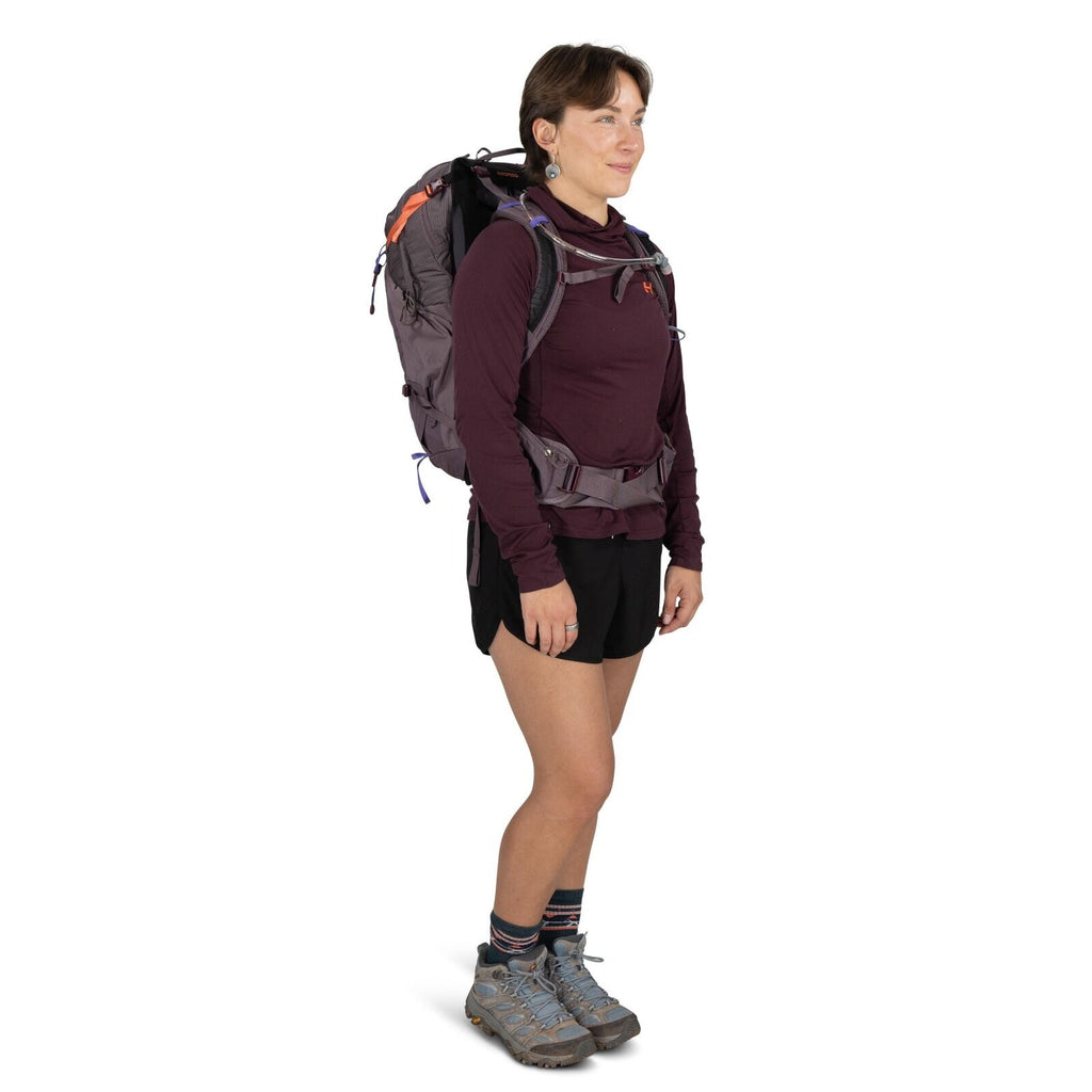 The image shows a person standing and dressed in outdoor hiking gear. They are wearing a maroon long-sleeve shirt with the logo 'HH' on the chest, and black shorts. On their back is a large, well-secured backpack with various straps and buckles. The perso
