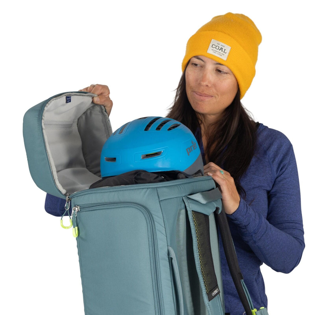 The image shows a person wearing a yellow beanie and a blue long-sleeved shirt. They are holding a teal-colored backpack, which is open, revealing the interior. Inside the backpack, a blue protective helmet is visible, situated on top of other packed item