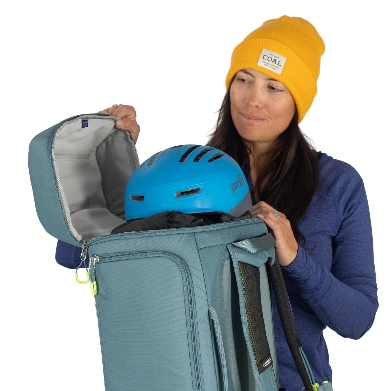 The image shows a person wearing a yellow beanie and a blue long-sleeved shirt. They are holding a teal-colored backpack, which is open, revealing the interior. Inside the backpack, a blue protective helmet is visible, situated on top of other packed item