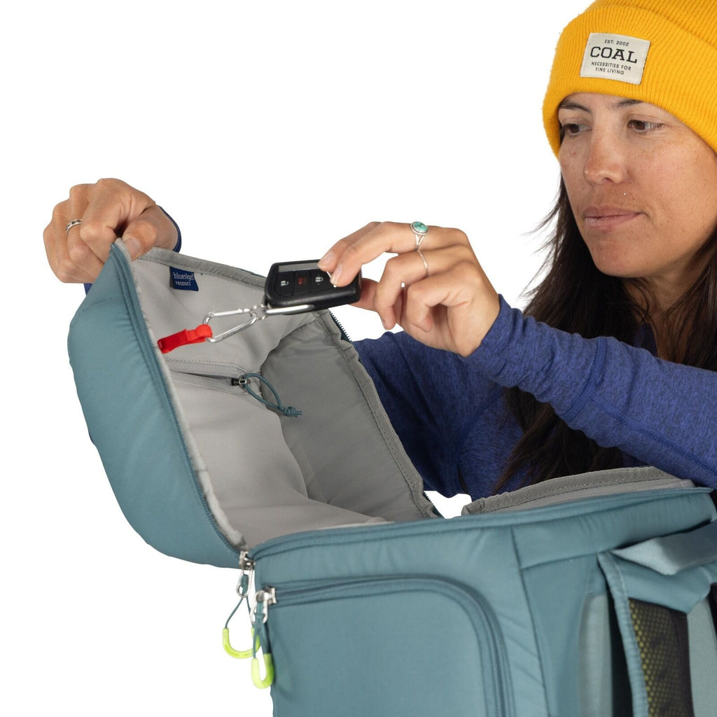 The image shows an individual wearing a yellow knit cap and a long-sleeved blue shirt, holding open a blue backpack with grey interior lining. They are demonstrating the use of a small digital scale with a red strap attached to the backpack, possibly to w