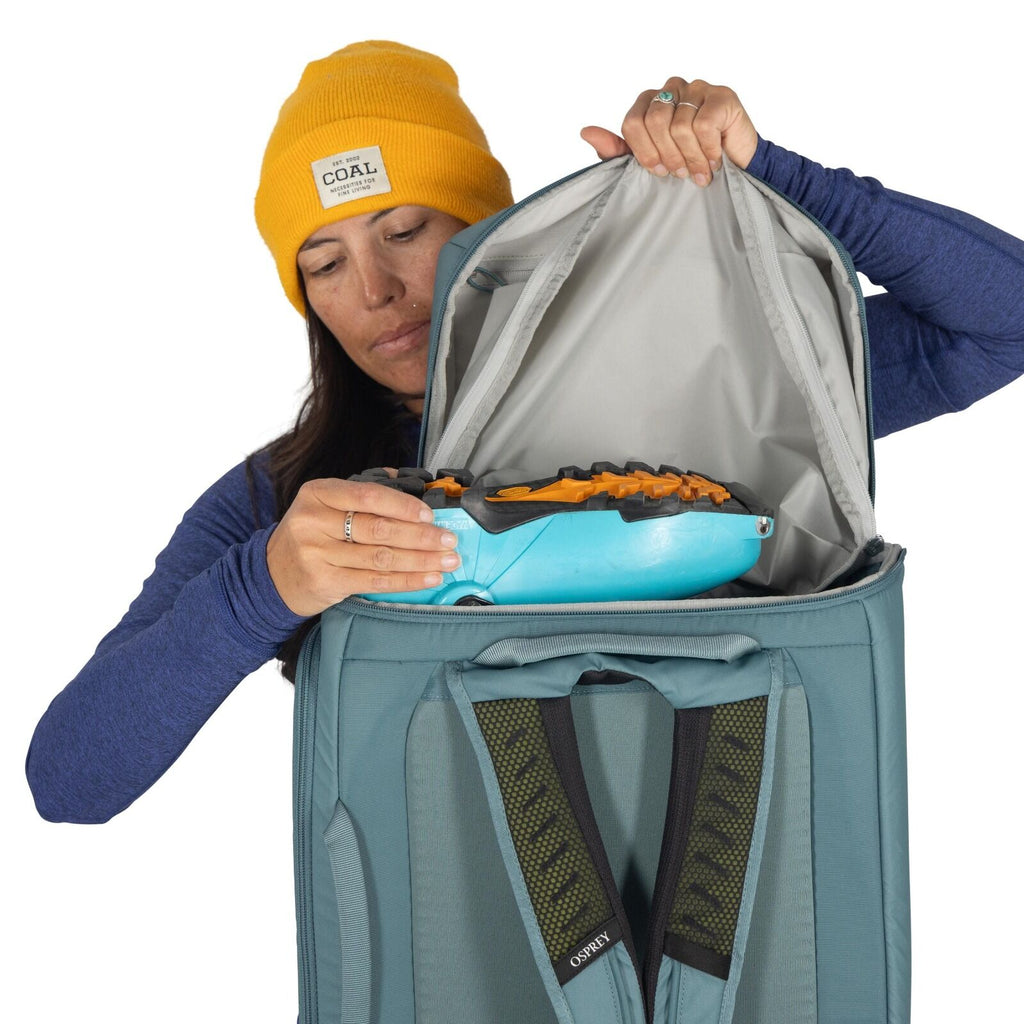The image depicts a person wearing a yellow beanie and a long-sleeved blue top, holding a blue and orange snowshoe in front of an open backpack. The backpack is gray with green straps and appears to be designed for travel or outdoor activities, suggesting