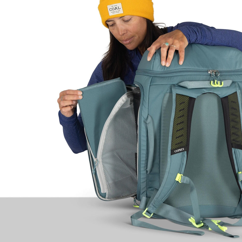 The image shows a person demonstrating the features of a blue Osprey backpack. The backpack is designed for travel, evident from its multiple compartments and padded straps for comfort. The person is holding open a zippered section, revealing an internal
