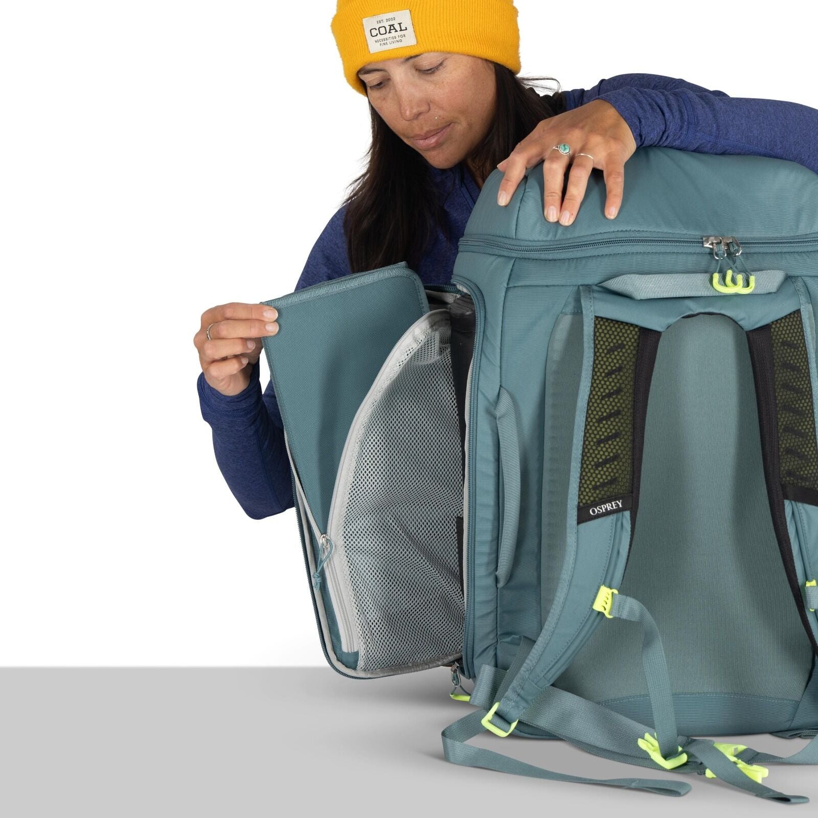 The image shows a person demonstrating the features of a blue Osprey backpack. The backpack is designed for travel, evident from its multiple compartments and padded straps for comfort. The person is holding open a zippered section, revealing an internal