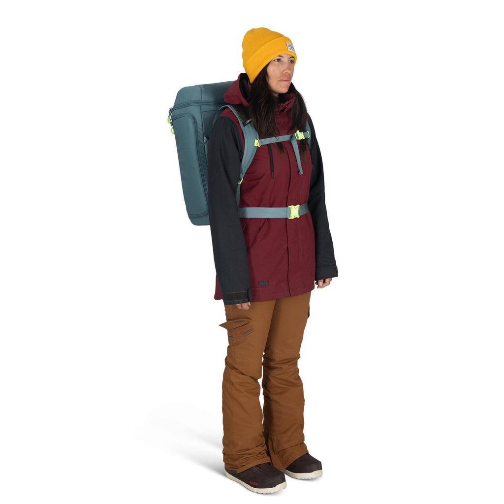 The image shows a person in a blue jacket holding open a black backpack, revealing its contents. The backpack appears to be a hiking or travel pack, made by the brand Osprey, which is visible on a label on the strap. Inside the backpack, there is a mesh c