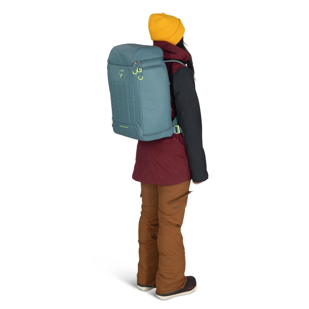 The image depicts a person standing sideways, showcasing a large backpack worn on their back. The backpack is light blue with some green accents and has a distinct square shape. The person is dressed warmly, wearing a mustard yellow beanie, a maroon jacke