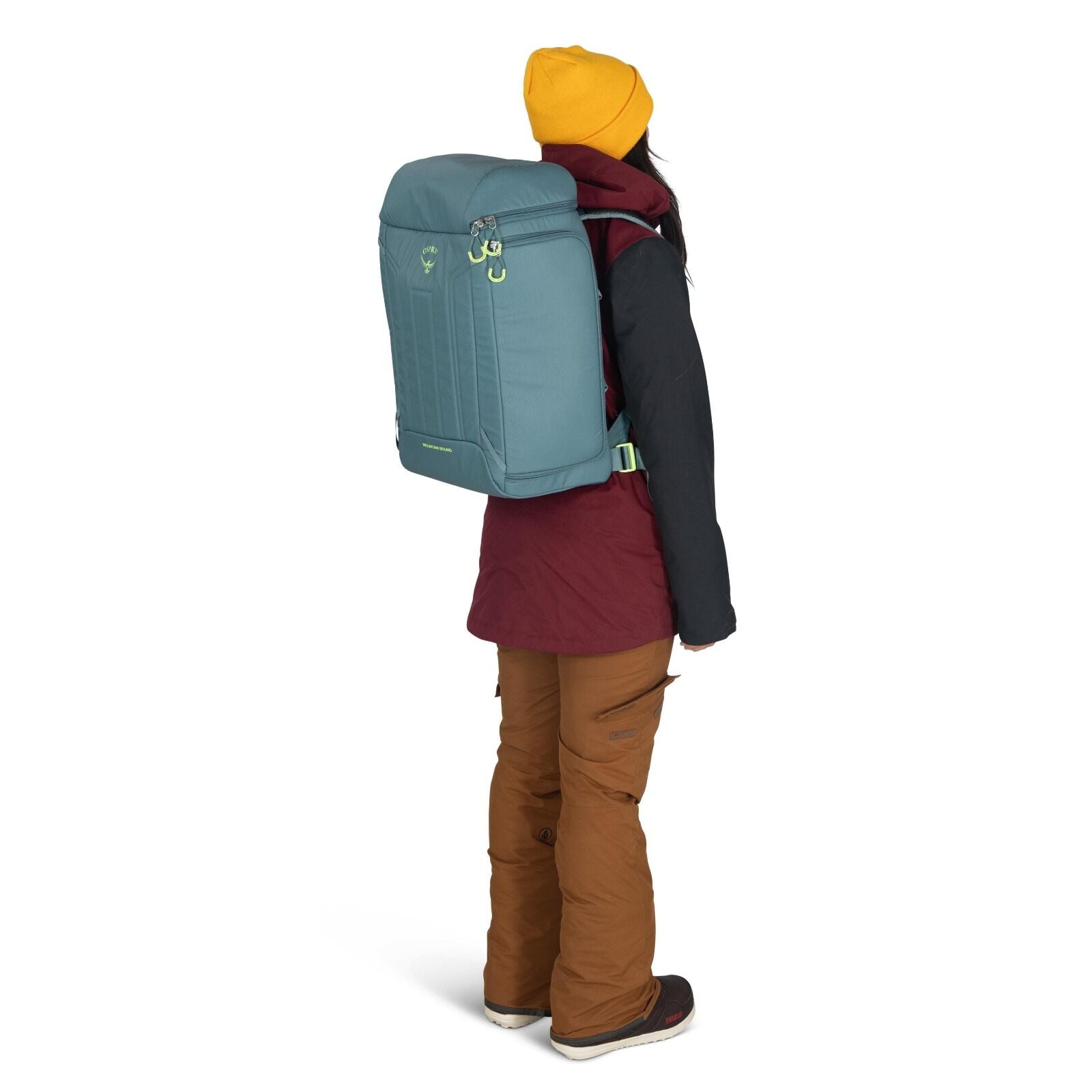 The image depicts a person standing sideways, showcasing a large backpack worn on their back. The backpack is light blue with some green accents and has a distinct square shape. The person is dressed warmly, wearing a mustard yellow beanie, a maroon jacke