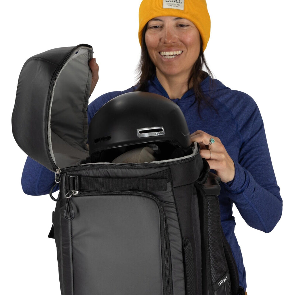 The image depicts a person wearing a blue long-sleeve shirt and a yellow hat. They are holding a large, dark gray backpack that appears to have multiple compartments and zippers. The top compartment of the backpack is open, revealing a black helmet inside