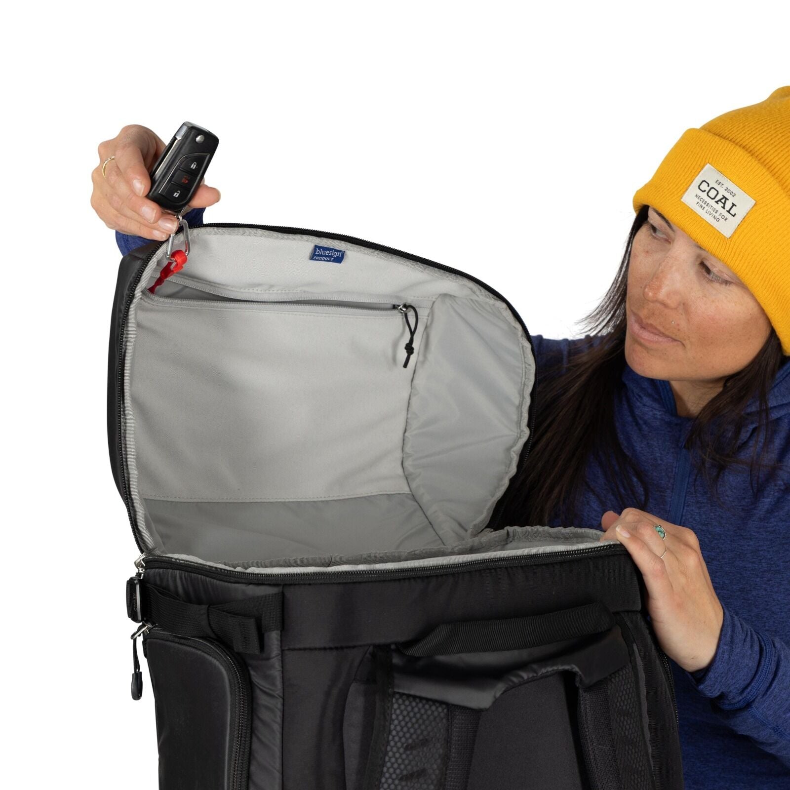 The image shows a person wearing a bright yellow beanie and a blue long-sleeve shirt, holding a black bag open. The interior of the bag is grey, and the person is pulling the bag's lid upwards to reveal its spacious compartment. In the person's left hand,