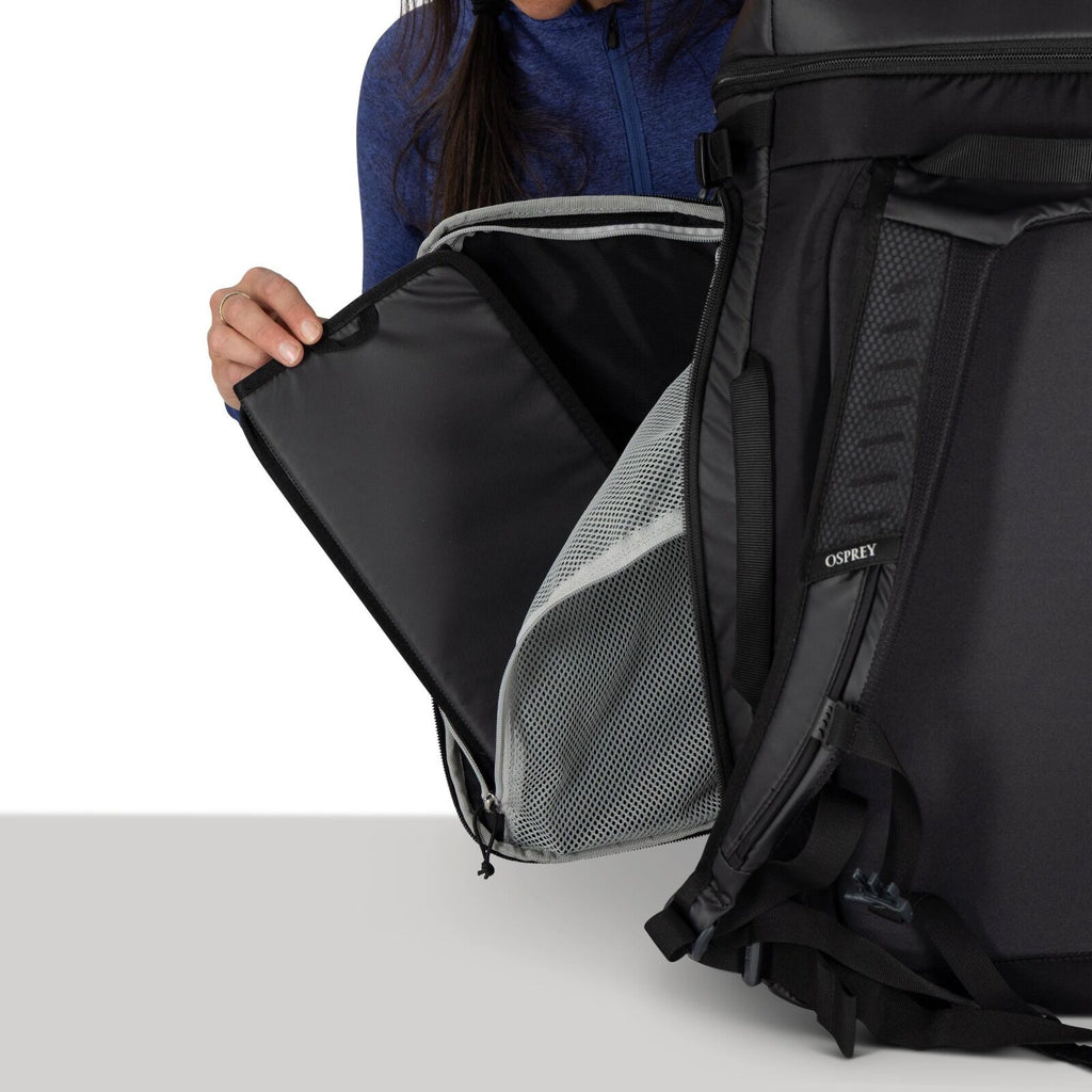 The image shows a person in a blue jacket holding open a black backpack, revealing its contents. The backpack appears to be a hiking or travel pack, made by the brand Osprey, which is visible on a label on the strap. Inside the backpack, there is a mesh c