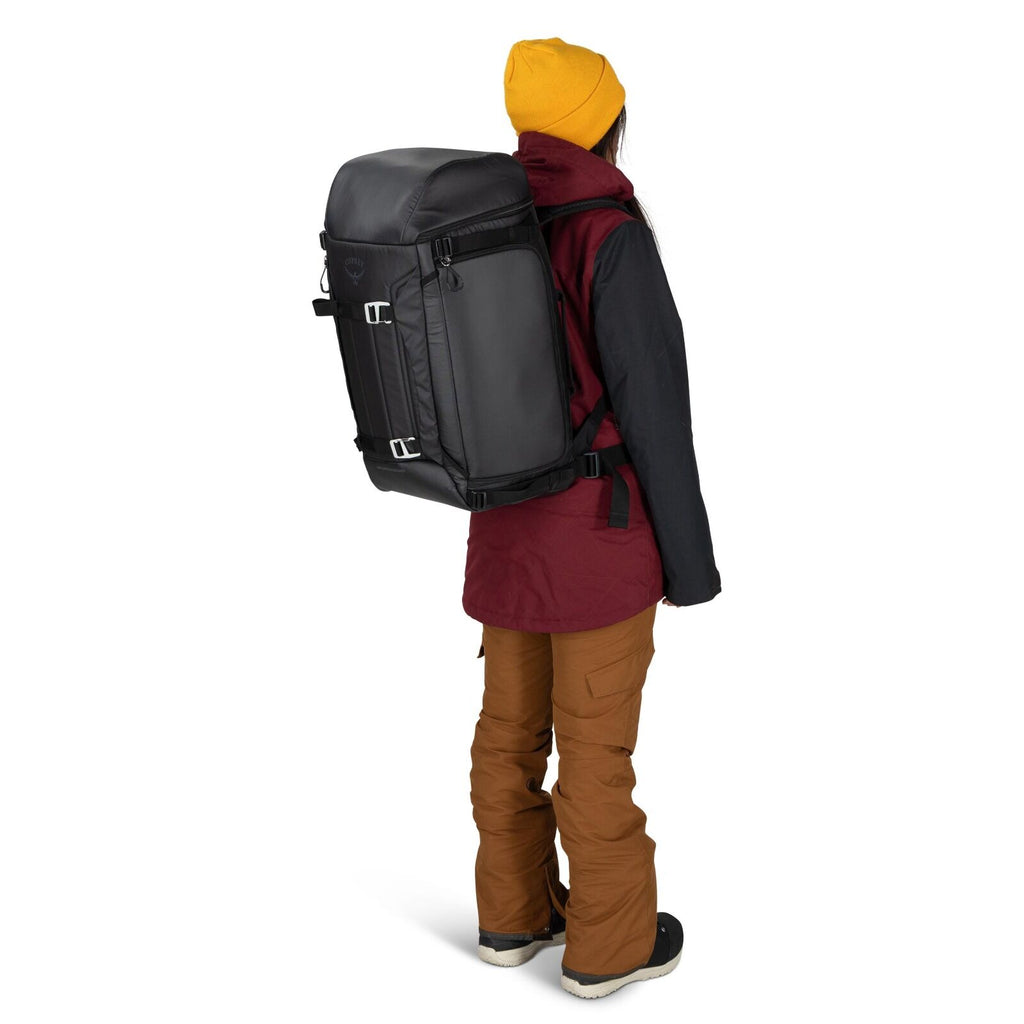 The image features a person standing with their back turned towards the camera, wearing outdoor winter attire. They have on a large black backpack that is securely fastened with straps and buckles. The individual is dressed in a maroon and dark gray winte