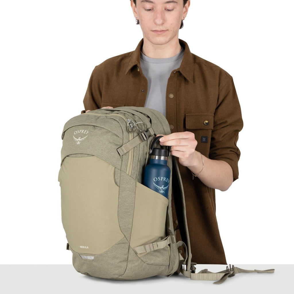 A person is holding a light-olive Osprey backpack with multiple zippered compartments and compression straps. They are placing a blue Osprey-branded water bottle into the stretch side pocket. The person wears a brown overshirt over a gray shirt, and their