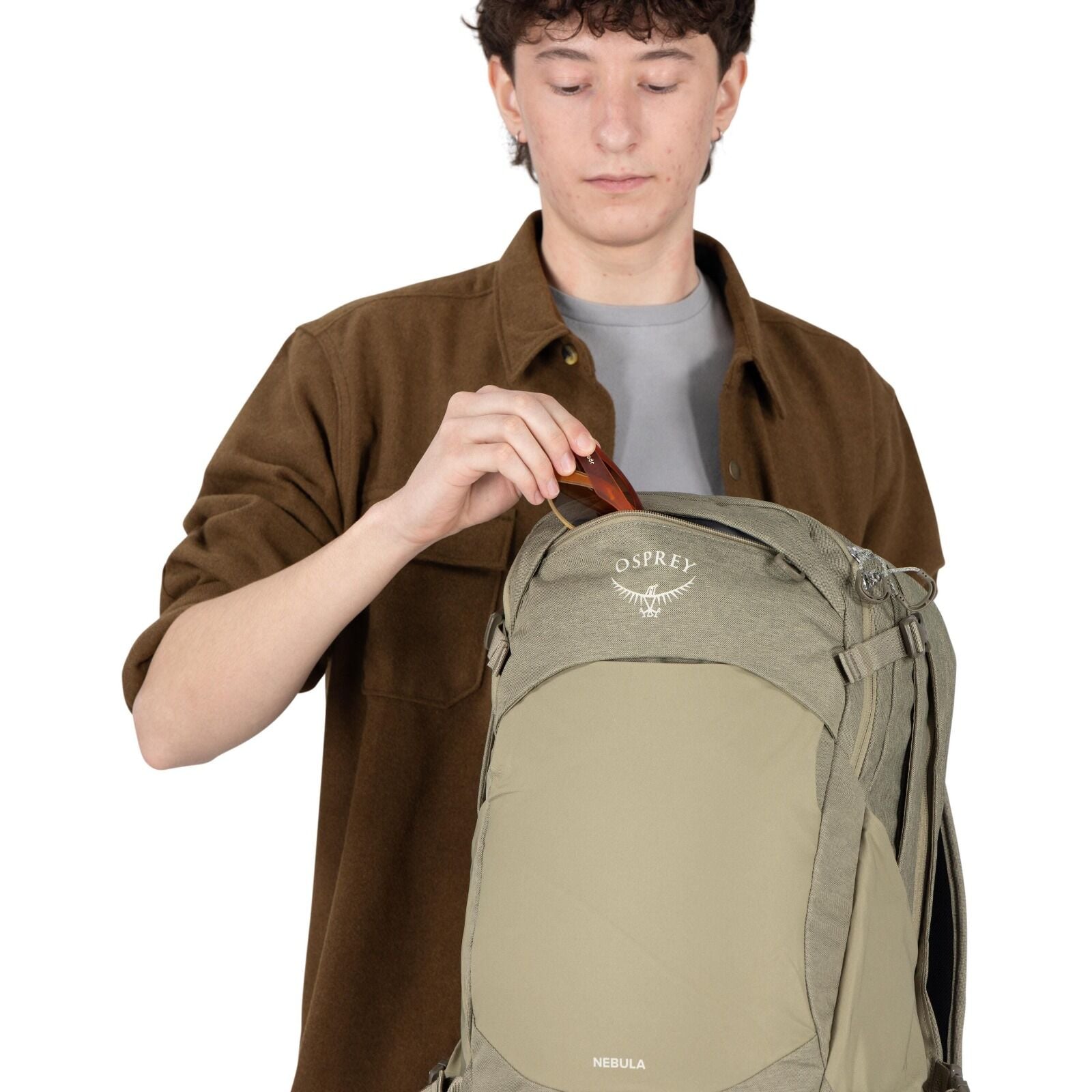 A person is placing a pair of sunglasses into the top pocket of an olive-green Osprey backpack. The model name "NEBULA" appears near the bottom front panel, and the Osprey bird logo is visible on the upper pocket. The person wears a brown overshirt over a