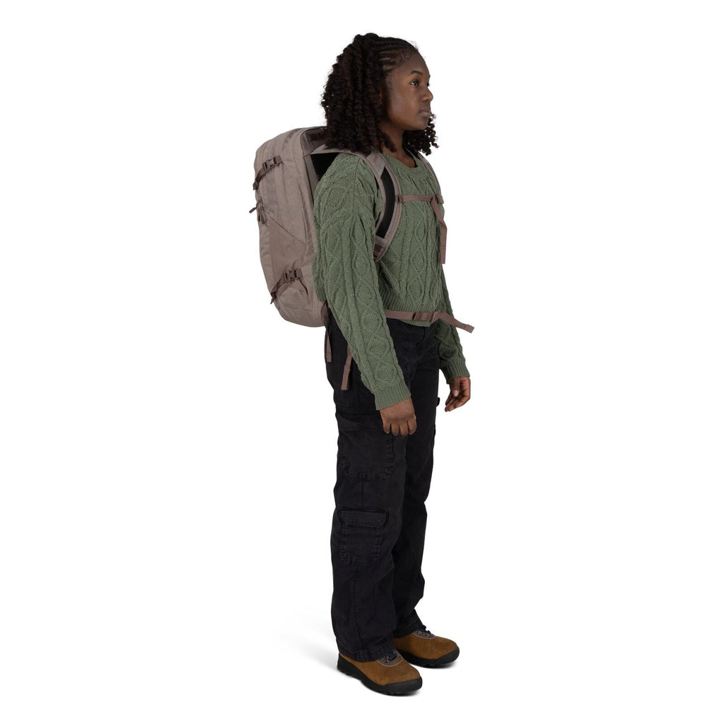A person stands in profile wearing outdoor clothing and a large beige backpack with chest and waist straps. They have on a green cable_knit sweater, black cargo pants, and brown hiking shoes with black soles. The backpack features side compression straps