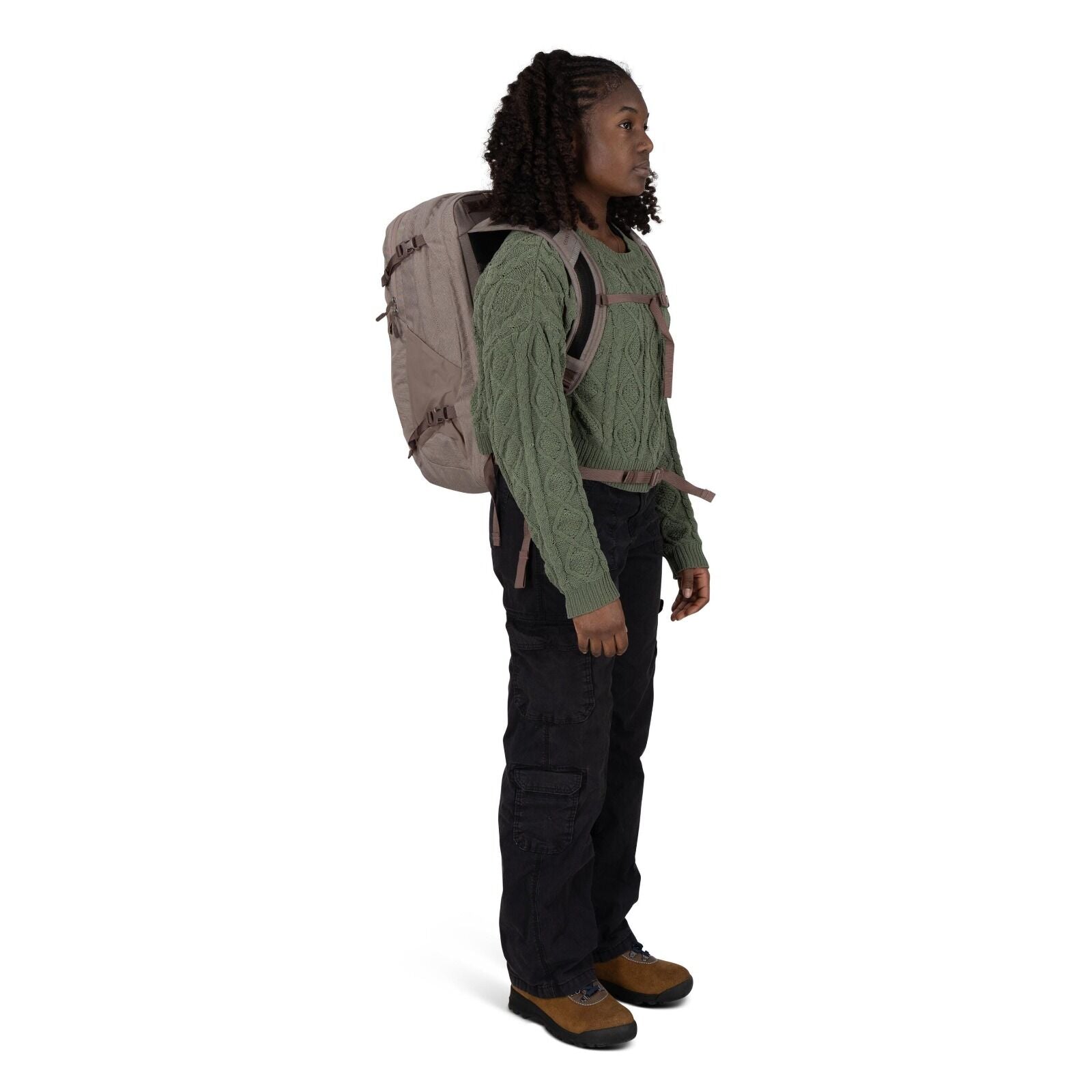 A person stands in profile wearing outdoor clothing and a large beige backpack with chest and waist straps. They have on a green cable_knit sweater, black cargo pants, and brown hiking shoes with black soles. The backpack features side compression straps