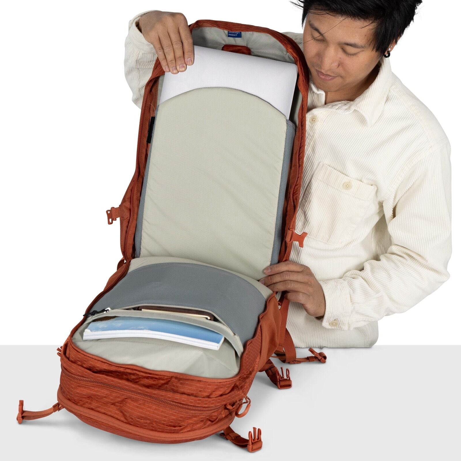 The image shows a person opening an orange backpack, revealing its spacious interior. Inside the backpack, a padded compartment holds a laptop, and in front of this, there is a large, shallow compartment with documents or folders. The interior of the back