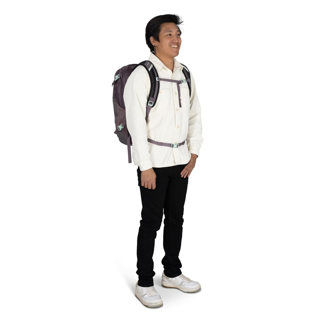 The image shows a person standing and dressed casually, with a purple backpack strapped securely. They are wearing a cream-colored button-up shirt and black pants, paired with white sneakers that have noticeable wear marks. The backpack features black str