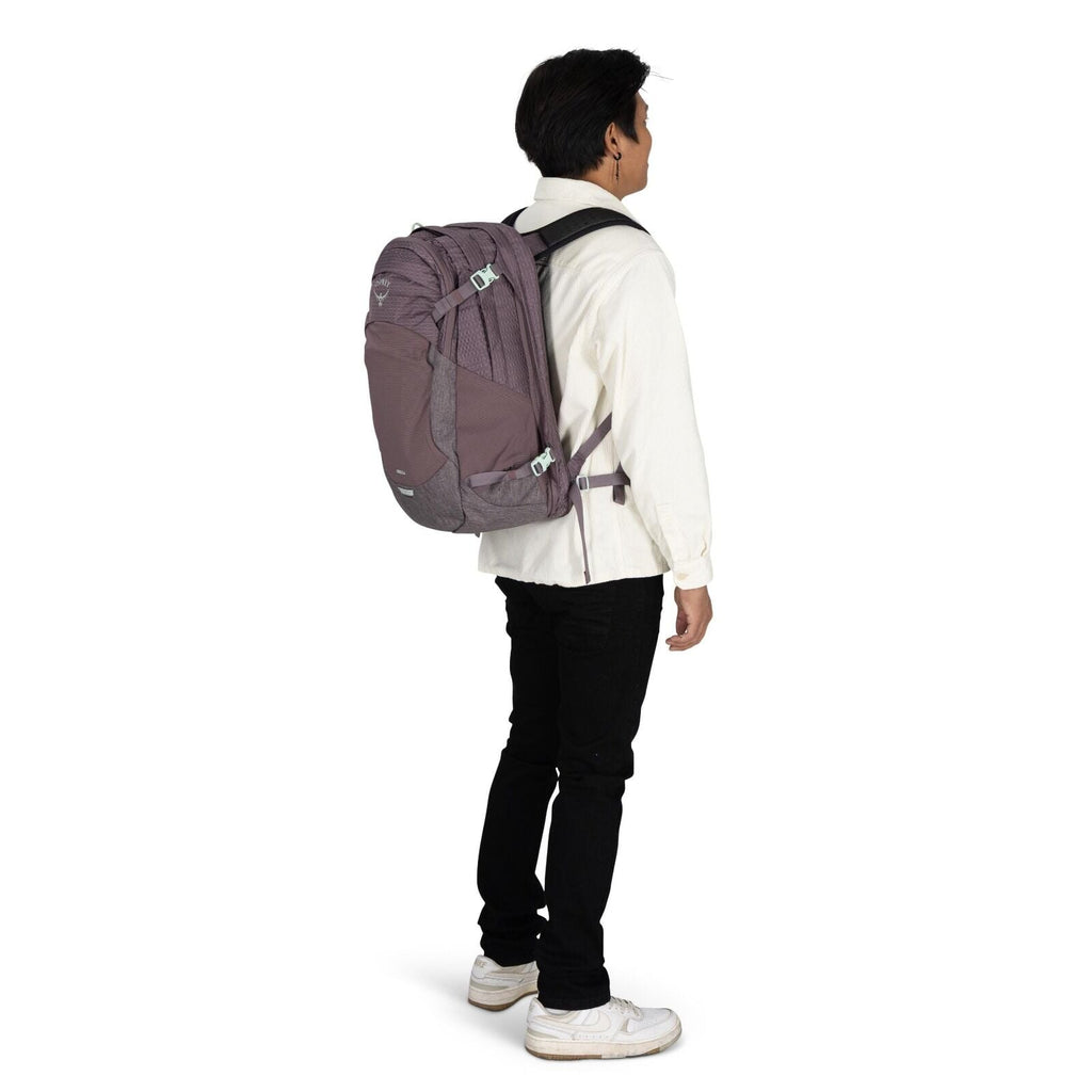 The image depicts a man standing in profile wearing a light purple Osprey backpack. He is dressed in a casual outfit consisting of a white button-up shirt, black pants, and white sneakers. The backpack appears to be spacious and suitable for travel or hik