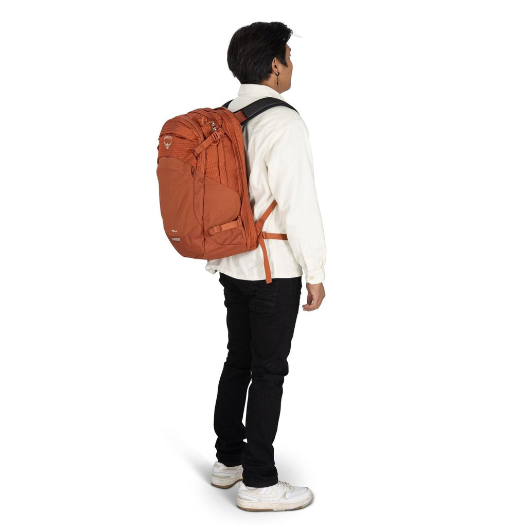 The image shows a person facing sideways, wearing an orange Osprey backpack. The backpack has multiple compartments and features a logo with the word "Osprey" and a graphic above it. The person is dressed in a white button-up shirt, black pants, and white