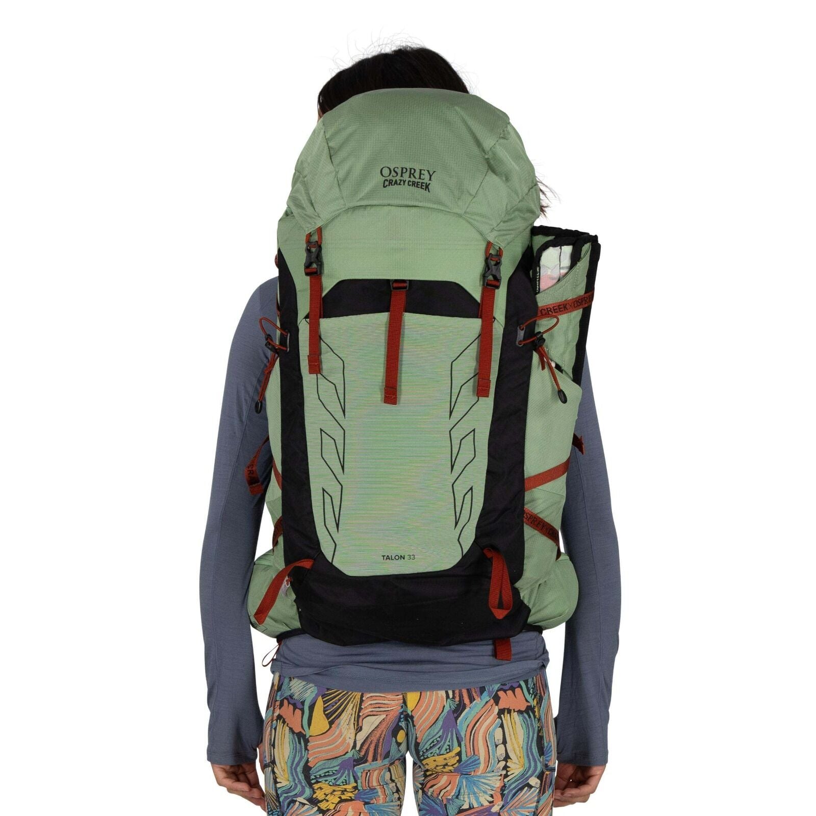 A person wearing a green Osprey / Crazy Creek Talon 33 backpack, featuring black accents and red straps, against a white background.