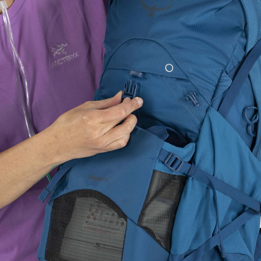 A person in a purple shirt adjusts a blue backpack strap. The backpack features multiple compartments and mesh details, conveying a functional, outdoor theme.