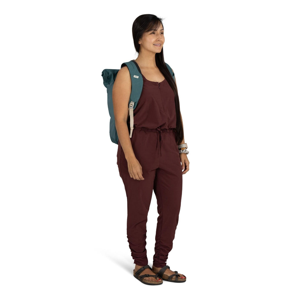 The image features a woman standing and dressed in a casual maroon jumpsuit with a drawstring at the waist. She is wearing black sandals and carries a teal backpack. Her long, dark hair is styled straight and falls over her shoulder. Additionally, she has
