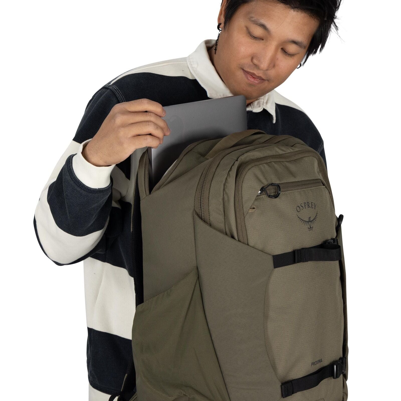 The image shows a person wearing a striped, long-sleeved shirt with wide black and white stripes, inserting a laptop into an Osprey backpack. The backpack is a neutral brown color and has the Osprey logo prominently displayed on the top front pocket. The