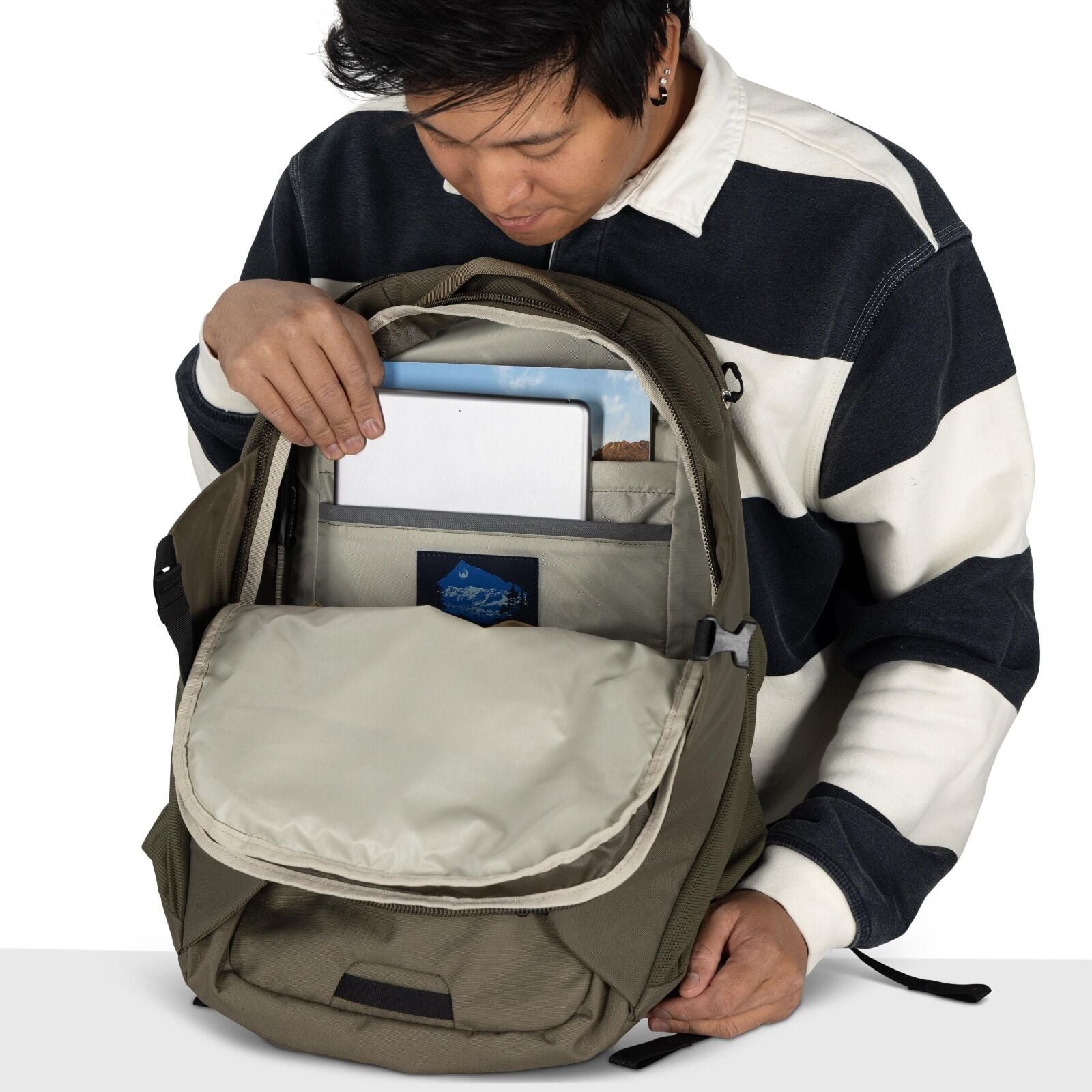 The image depicts a person holding open a backpack to reveal its contents. The backpack is olive green with a beige interior. Inside the backpack, there is a tablet in a dedicated pocket and a postcard featuring a mountainous landscape behind it. Below th