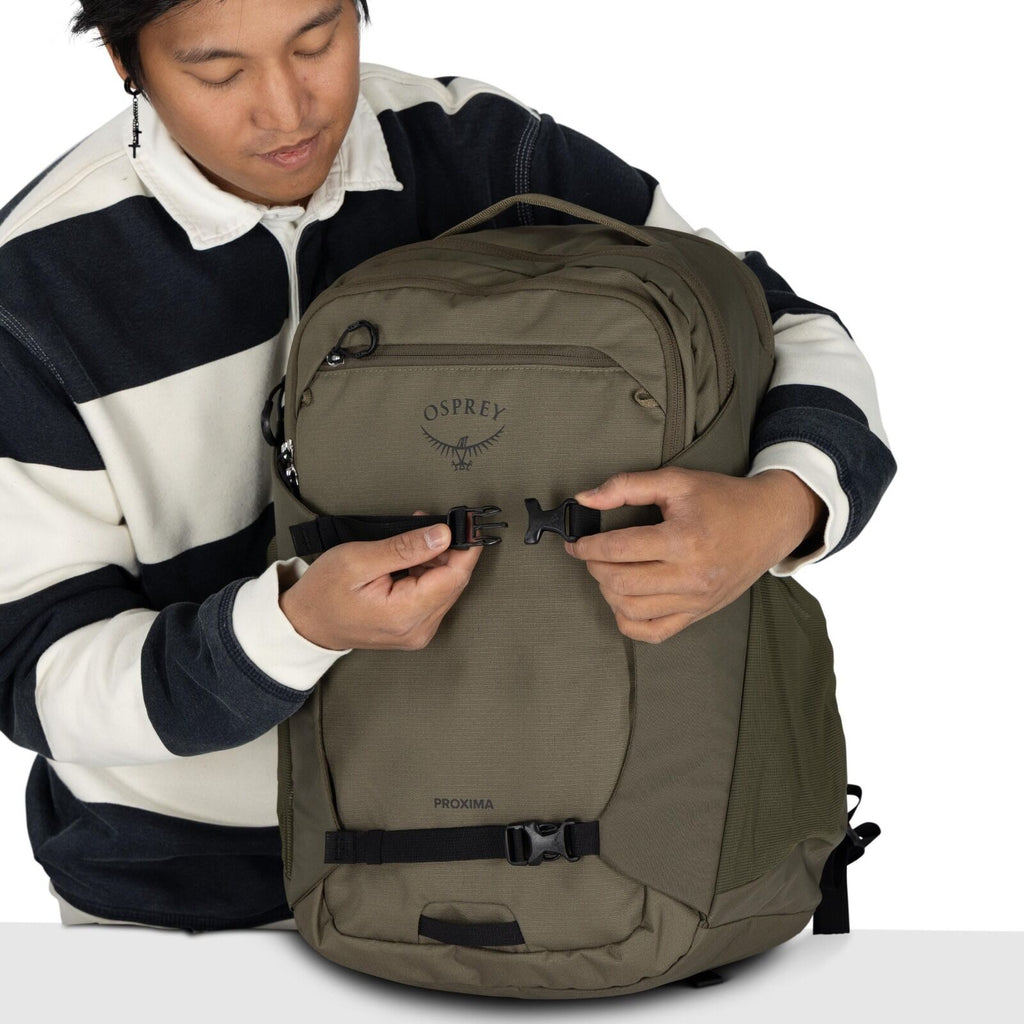 The image shows a person wearing a black and white striped sweater, holding a large backpack. The backpack is olive green and features the "Osprey" brand logo with an accompanying wing design prominently displayed on the front. The person is adjusting one