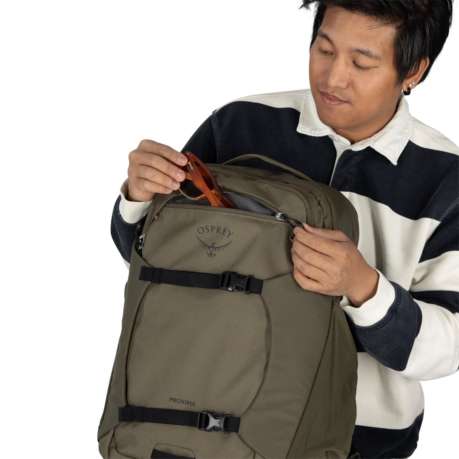 The image shows a person holding a khaki-colored Osprey backpack model "Proxima." The person is wearing a black and white striped long-sleeve shirt and is in the process of placing or retrieving a pair of brown sunglasses from the backpack's front pocket.