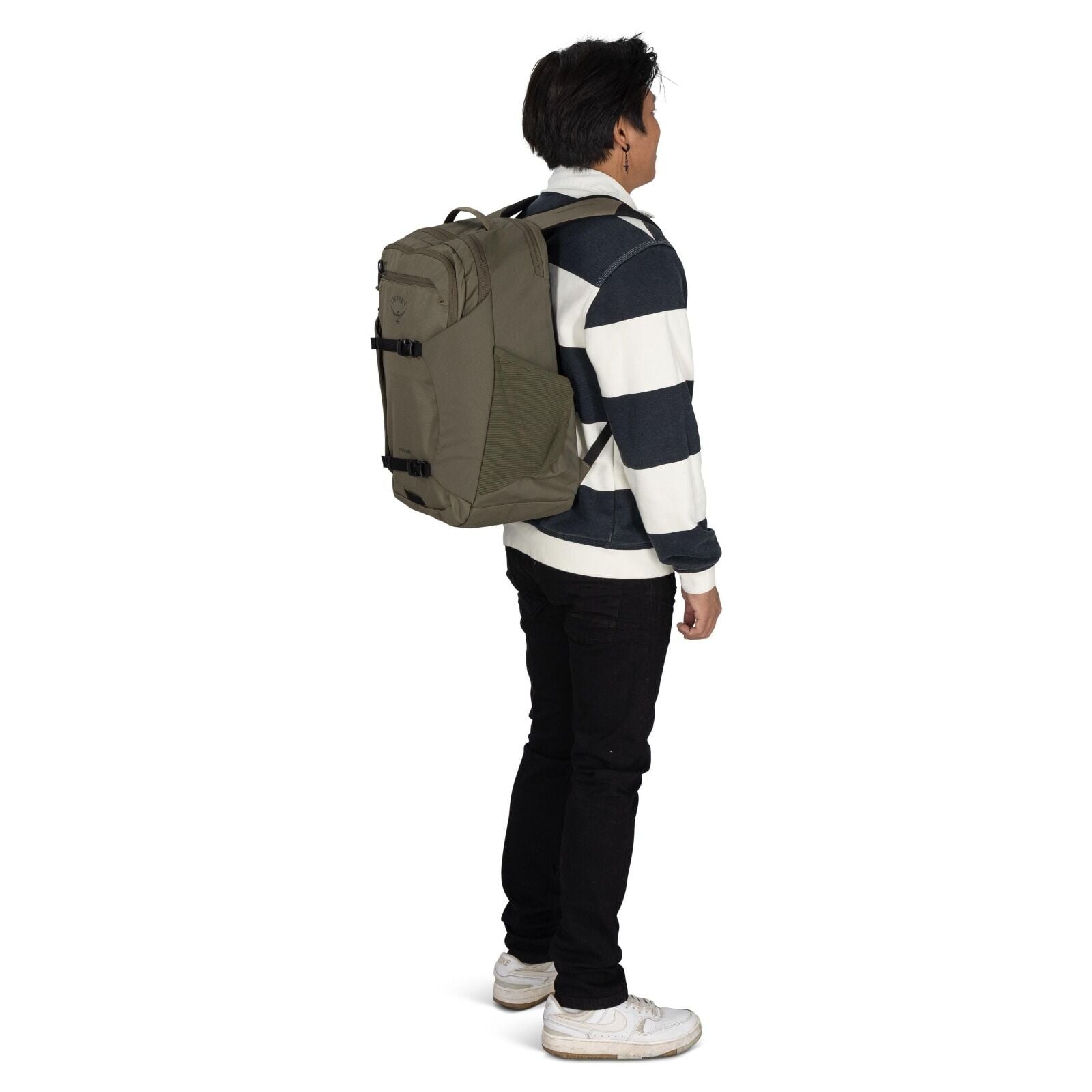 The image depicts a young man standing and facing slightly to the side. He is wearing a striped sweater with dark navy and white horizontal stripes, paired with black pants and white sneakers. On his back, he is carrying a large, olive-green backpack with
