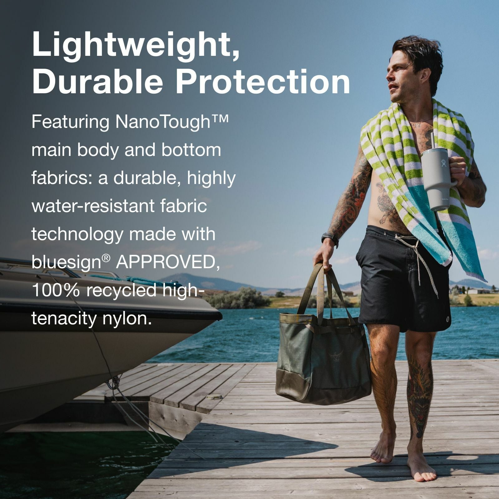 A man stands on a dock by the water, holding a tote bag and a drink, showcasing lightweight, water-resistant outdoor apparel.