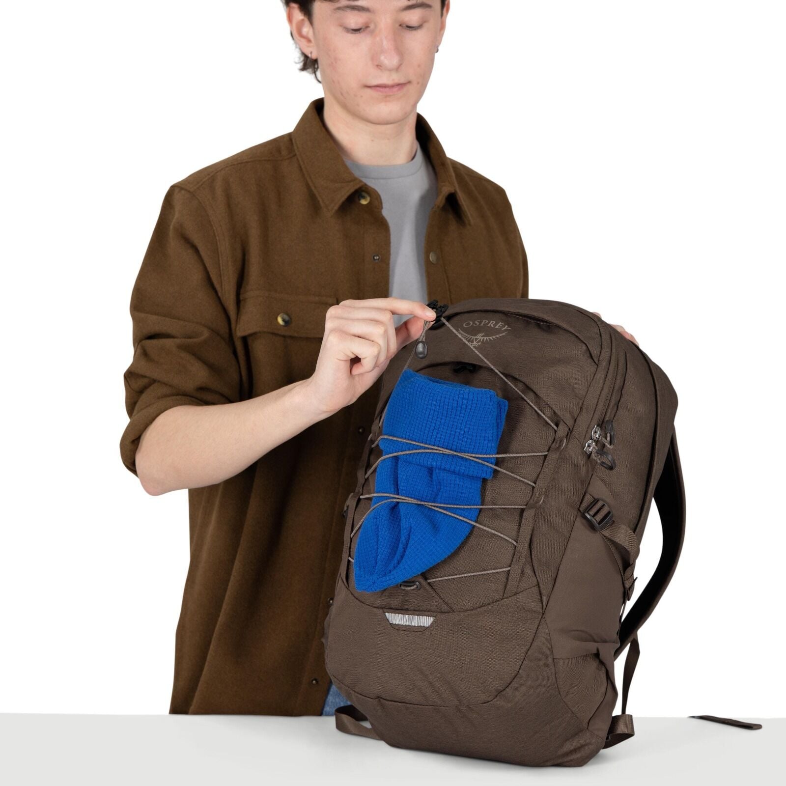 A person with a blurred face is standing behind a table, holding a cream-colored cap in one hand and resting the other hand on a large brown backpack. The backpack is an Osprey Quasar model with a front bungee cord system, side compression straps, and mul