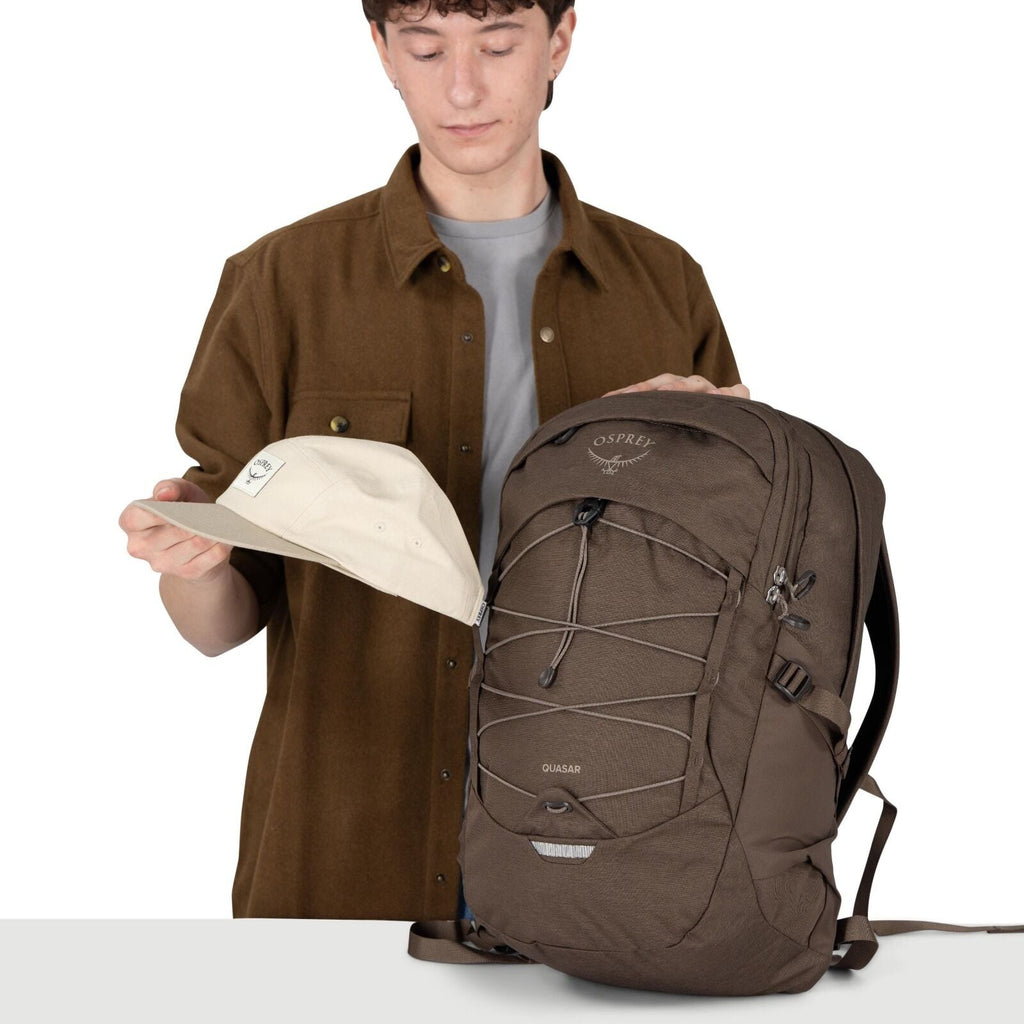 A person with a blurred face is standing behind a table, holding a cream-colored cap in one hand and resting the other hand on a large brown backpack. The backpack is an Osprey Quasar model with a front bungee cord system, side compression straps, and mul