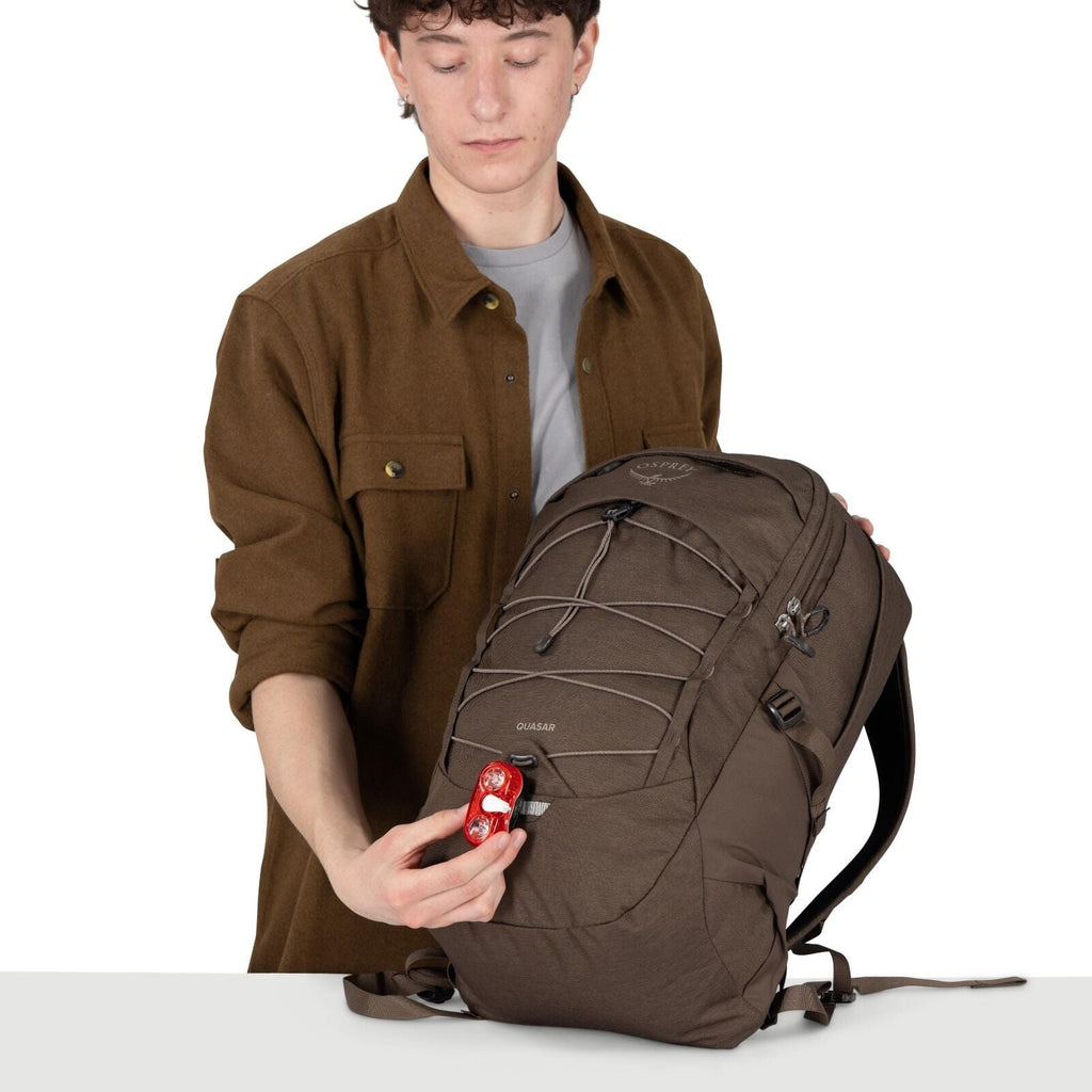 A person holds a brown Osprey Quasar backpack on a white surface. They're attaching a small red bike light to a loop on the front of the pack. The backpack features front bungee cords, multiple zippered compartments, and padded shoulder straps. The person