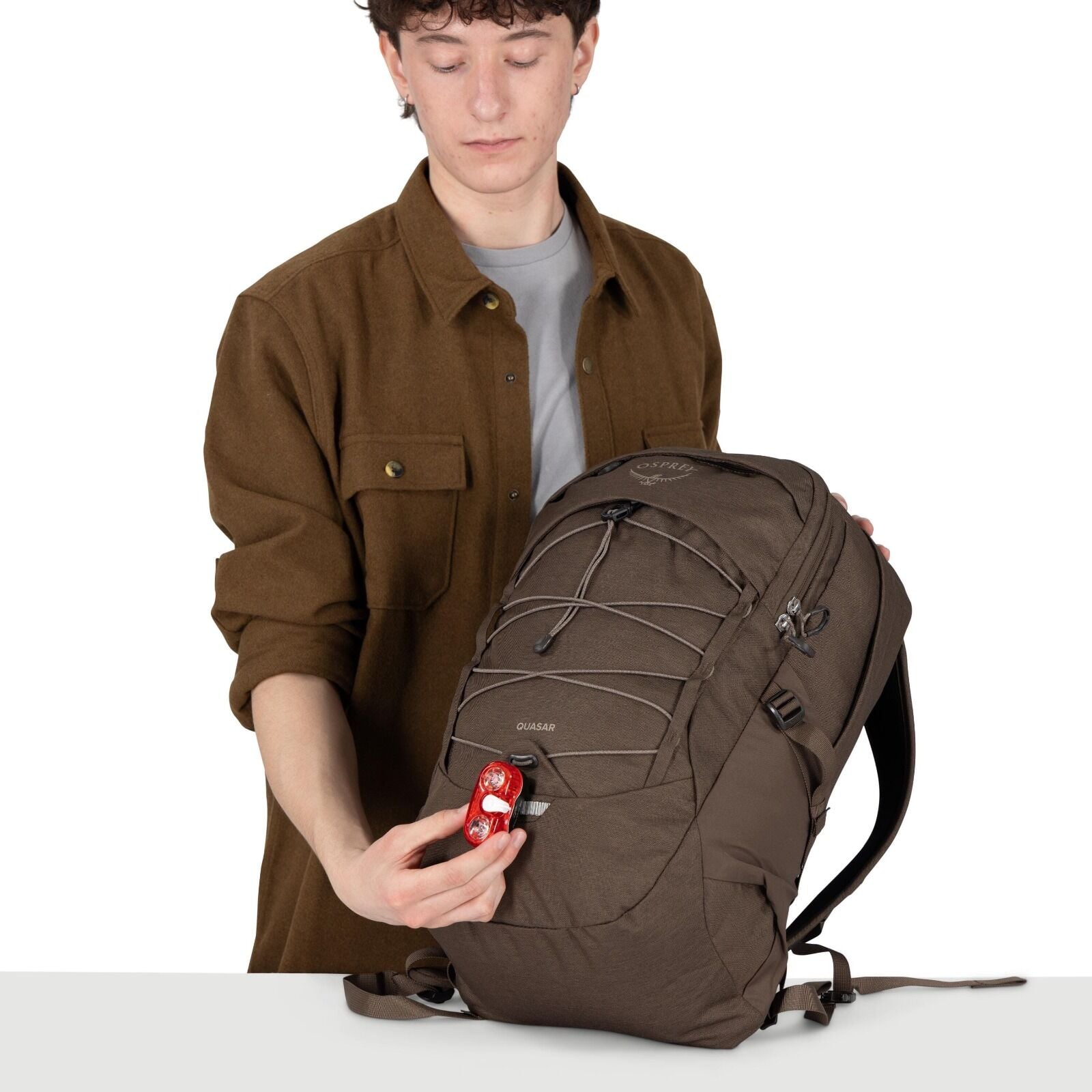 A person holds a brown Osprey Quasar backpack on a white surface. They're attaching a small red bike light to a loop on the front of the pack. The backpack features front bungee cords, multiple zippered compartments, and padded shoulder straps. The person