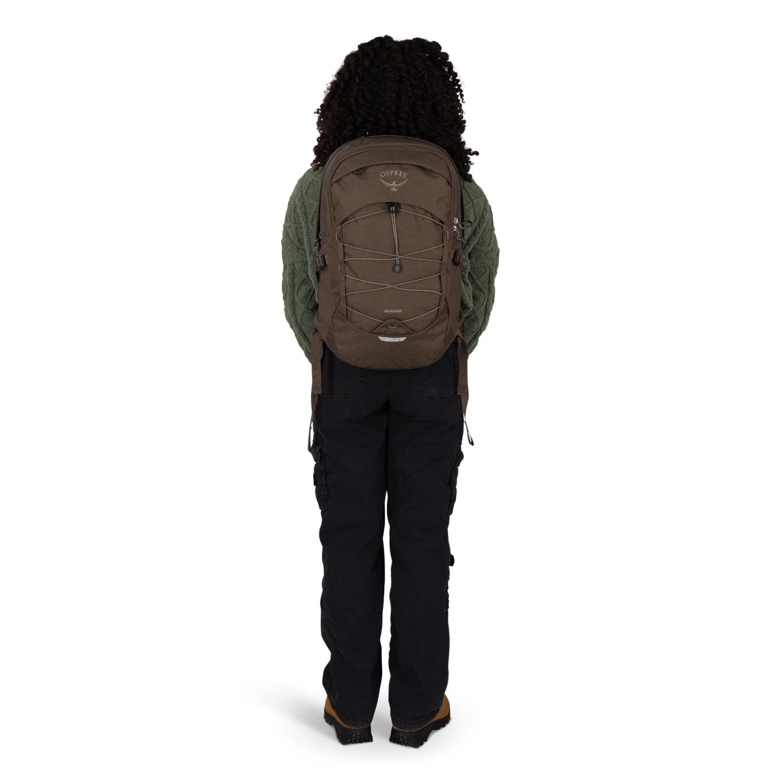 A person is standing facing away from the camera, wearing a brown Osprey backpack. The backpack has a front bungee cord system, side compression straps, and a small top pocket. They are dressed in a textured green sweater, black cargo-style pants, and bro