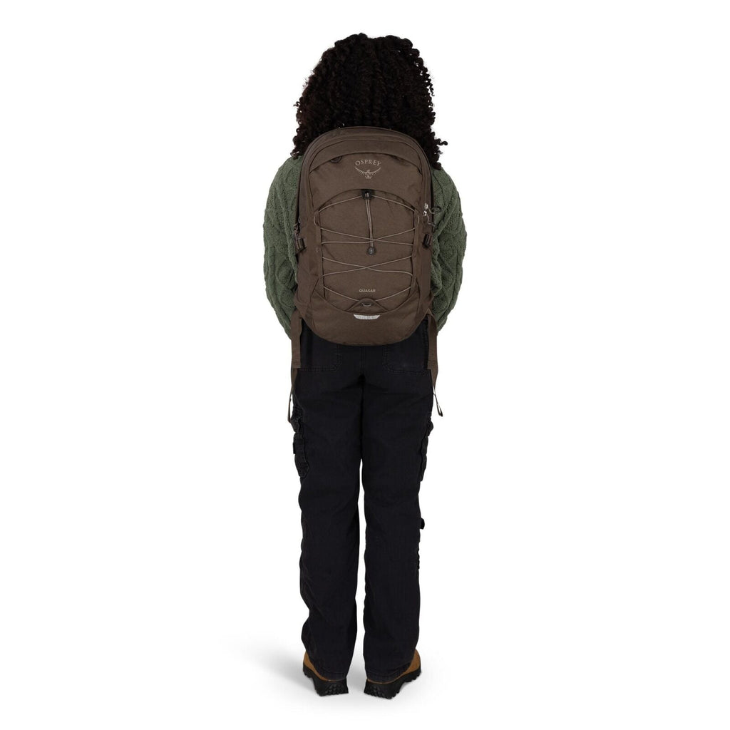 A person is standing facing away from the camera, wearing a brown Osprey backpack. The backpack has a front bungee cord system, side compression straps, and a small top pocket. They are dressed in a textured green sweater, black cargo-style pants, and bro