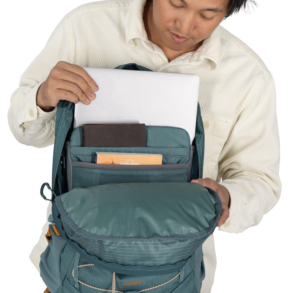 The image depicts a person wearing a cream-colored, corduroy shirt, opening a teal Quasar brand backpack to reveal its contents. Inside the backpack, a laptop is seen being pulled out from a dedicated laptop sleeve. The backpack also contains several item