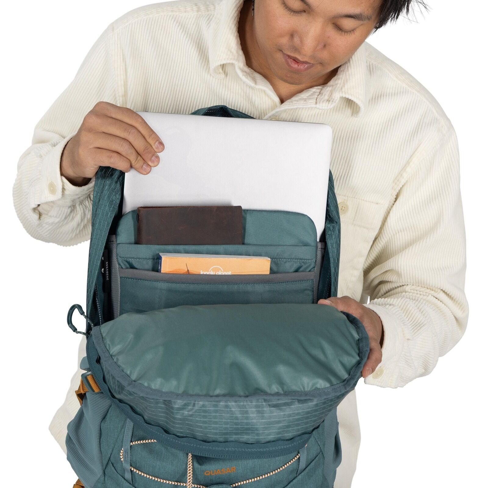 The image depicts a person wearing a cream-colored, corduroy shirt, opening a teal Quasar brand backpack to reveal its contents. Inside the backpack, a laptop is seen being pulled out from a dedicated laptop sleeve. The backpack also contains several item