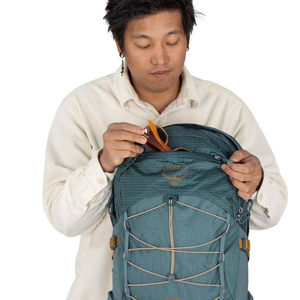 The image depicts a person wearing a beige, corduroy button-up shirt. They are holding a teal Osprey backpack, prominently displaying the brand's logo. The backpack features multiple zippered compartments and orange accents, such as straps and cords, enha