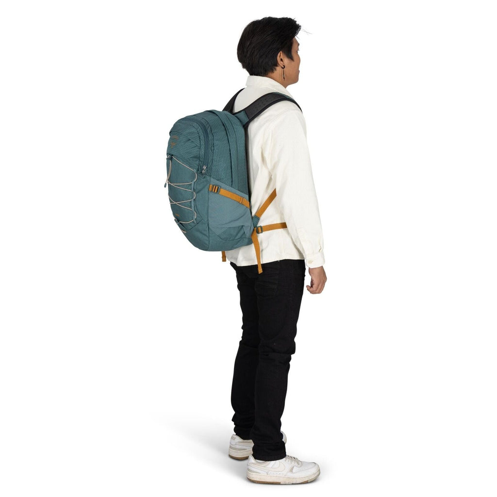 The image depicts a man standing sideways, showcasing a large teal backpack on his back. He is wearing a white button-up shirt, black jeans, and white sneakers. The backpack features a distinctive design with orange straps and a bungee cord system on its