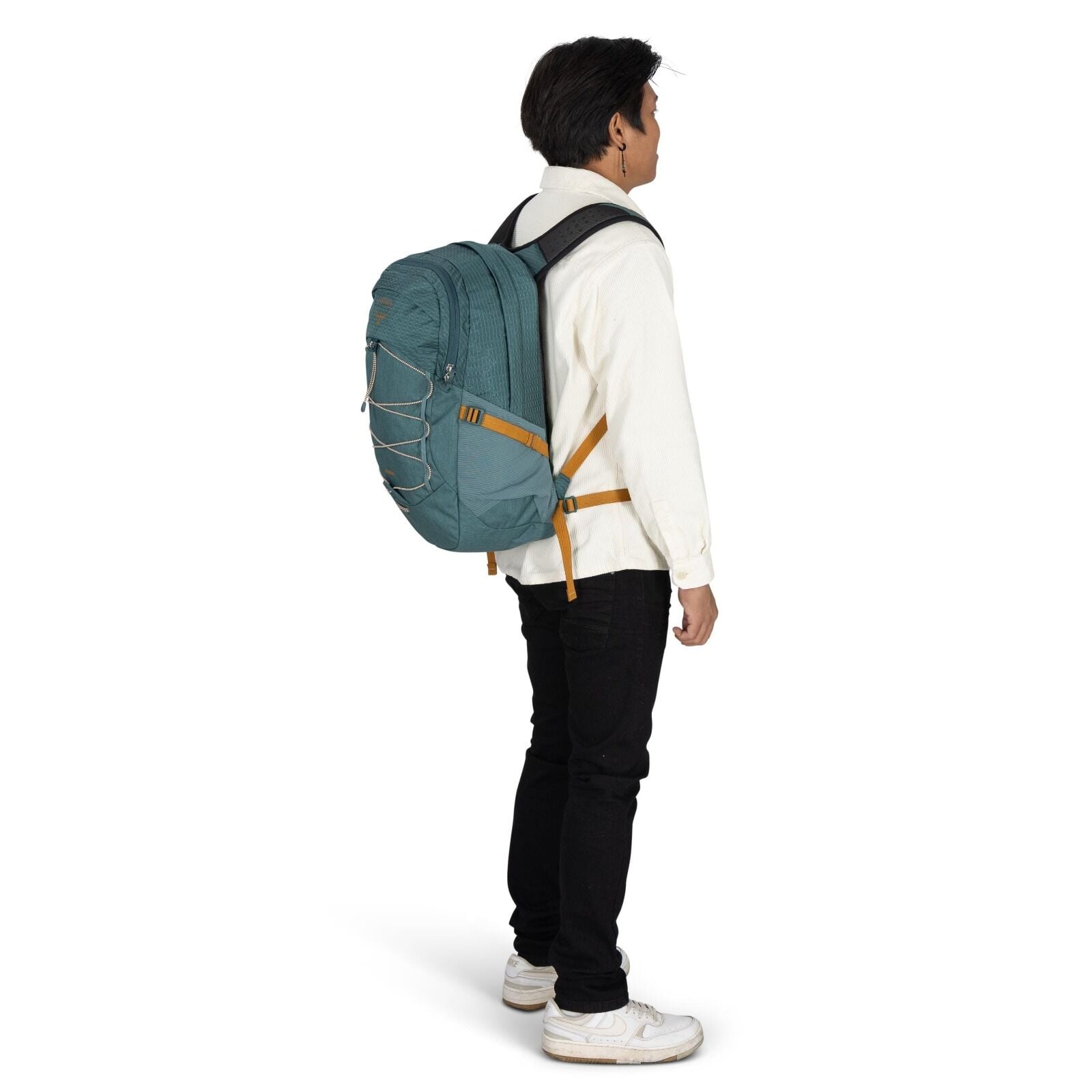 The image depicts a man standing sideways, showcasing a large teal backpack on his back. He is wearing a white button-up shirt, black jeans, and white sneakers. The backpack features a distinctive design with orange straps and a bungee cord system on its