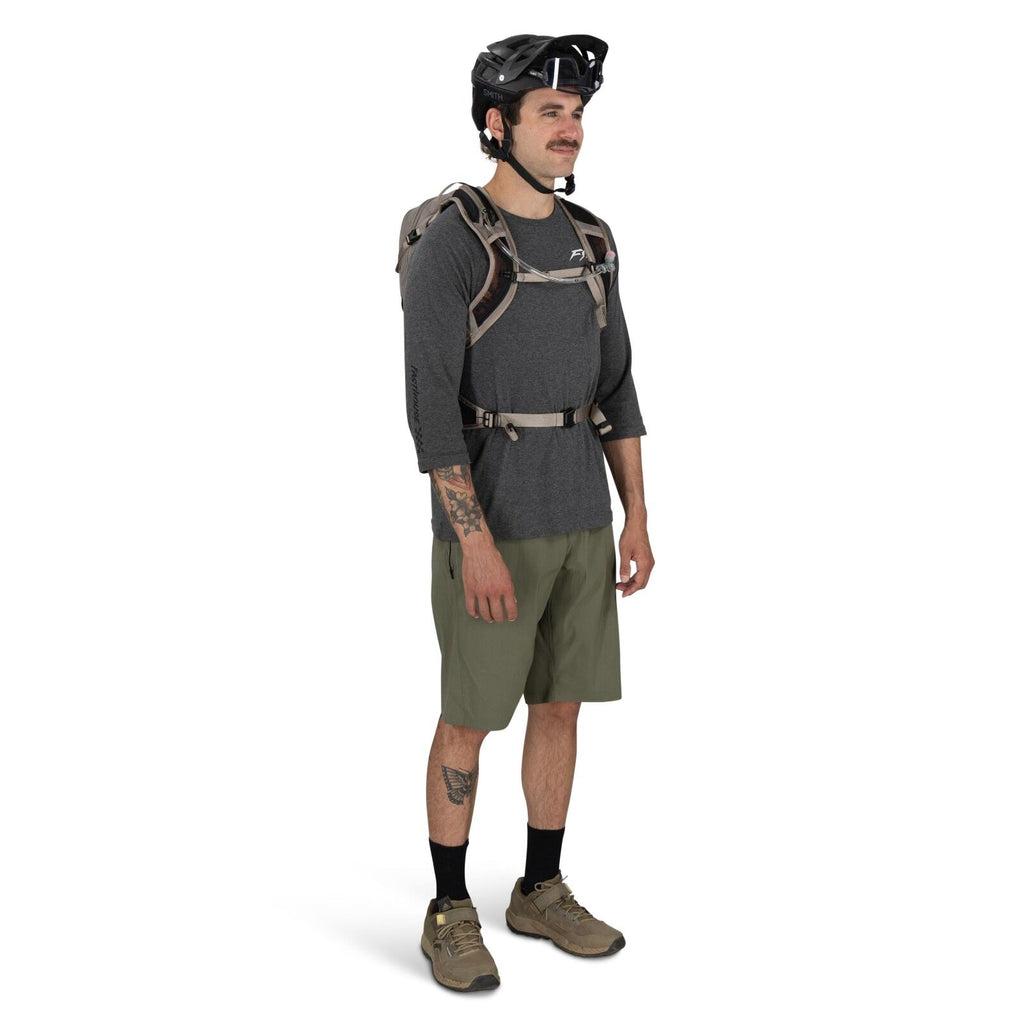 The image features an individual dressed in casual outdoor attire. The person is wearing a gray long-sleeved shirt with a logo on the chest area and olive green shorts. They have a tattoo on their left forearm and a tattoo on their left calf. The individu