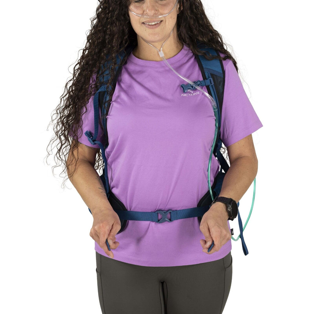 A woman with long, curly hair wears a purple T-shirt, blue backpack, and oxygen tubing. She appears ready for hiking and is smiling slightly.