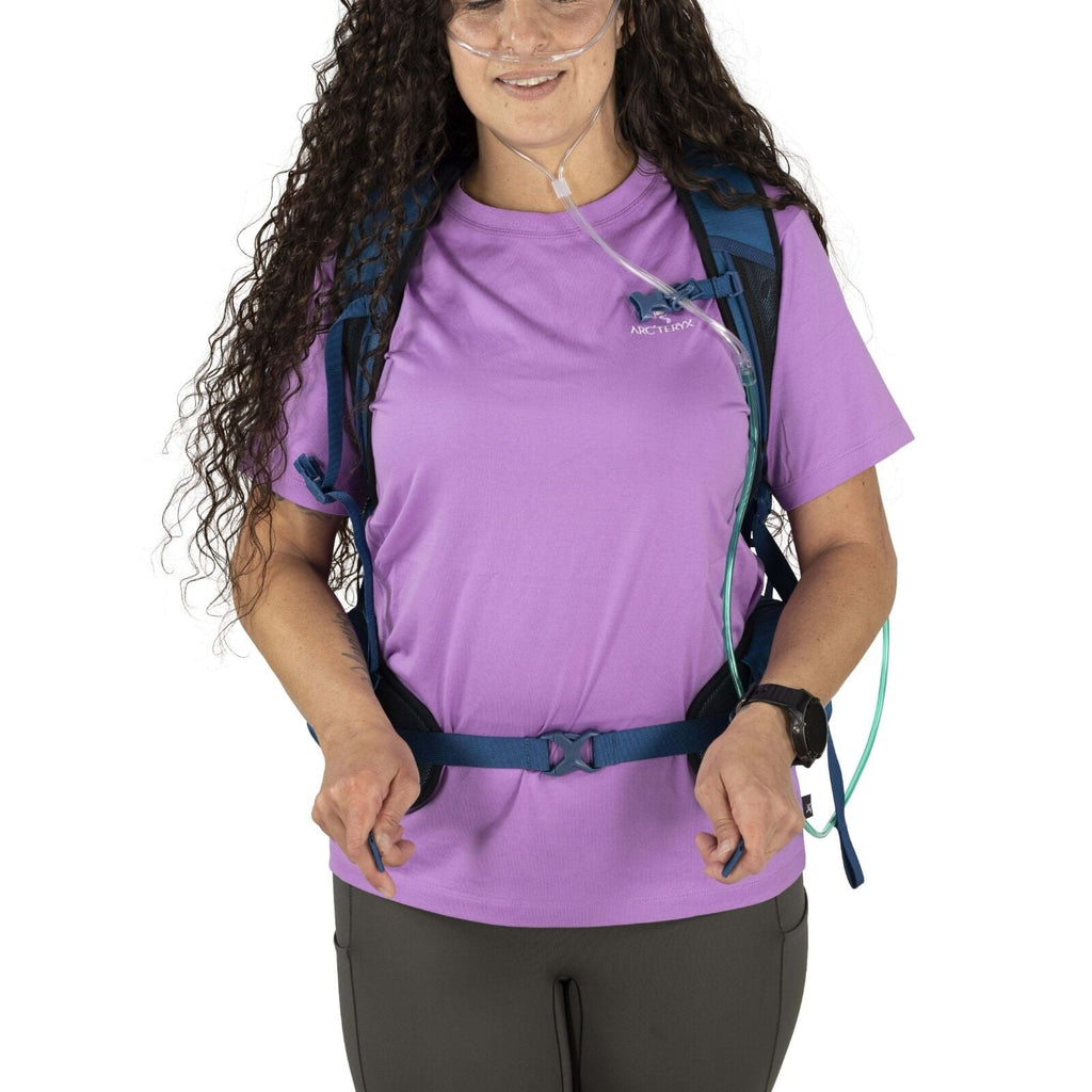 A woman with long, curly hair wears a purple T-shirt, blue backpack, and oxygen tubing. She appears ready for hiking and is smiling slightly.