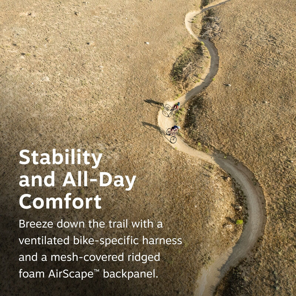 The image depicts two mountain bikers riding along a winding dirt trail on a grassy hillside. The trail snakes back and forth, creating a dynamic zigzag pattern as it traverses the natural landscape. The scene is captured from an aerial perspective, offer