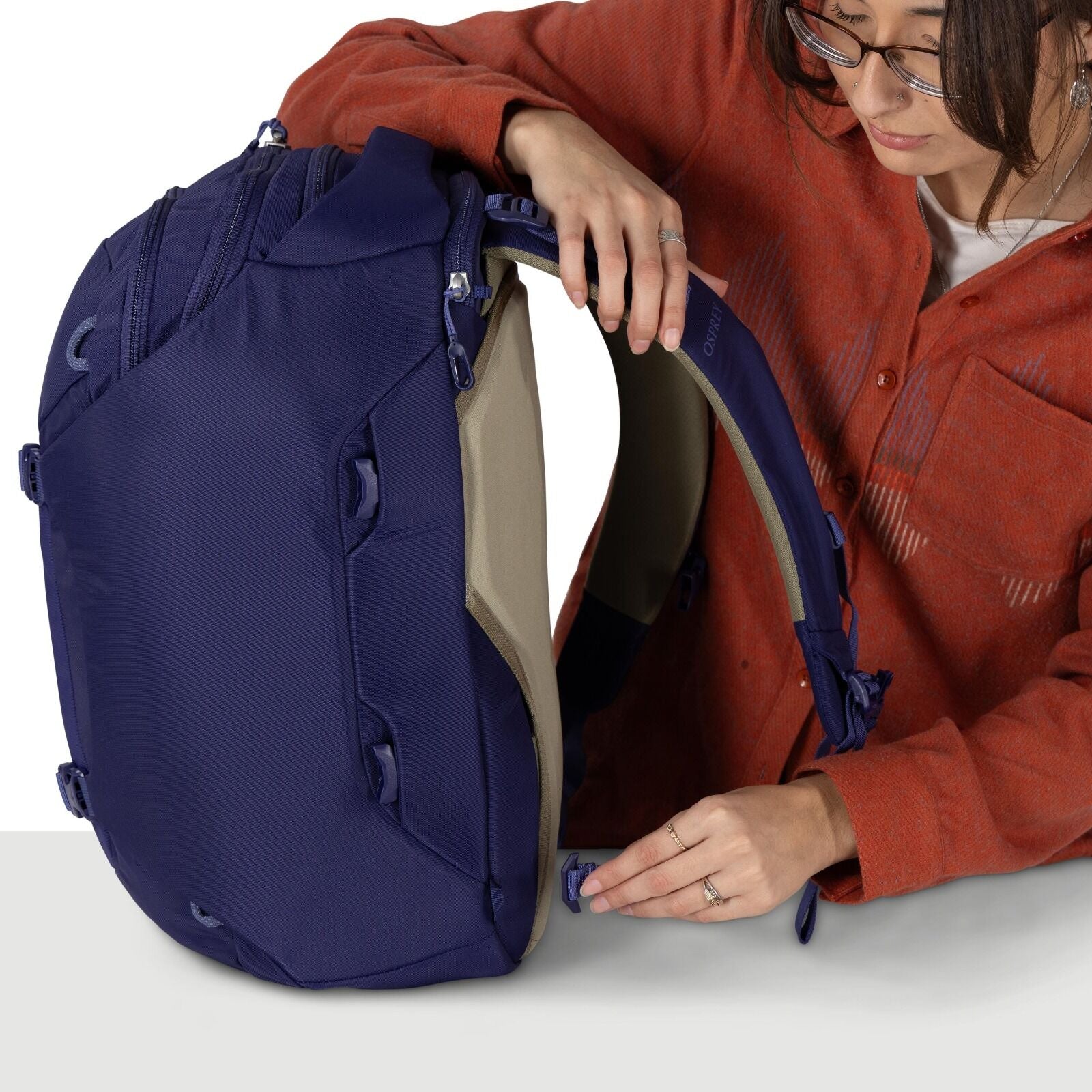 A person wearing a red flannel shirt  is packing an rich purple Osprey backpack. They are adjusting a strap on the back of the pack. The scene is well lit with a plain white background.
